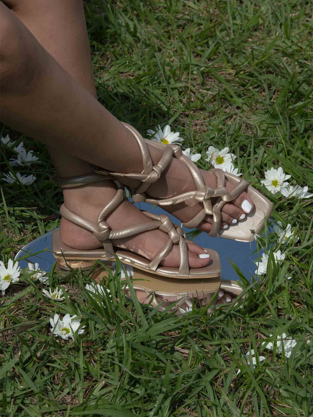 Detalle de los pies con la sandalia plana Duna dorada, capturando el brillo de los nudos artesanales y el mini tacón de madera bajo la luz natural