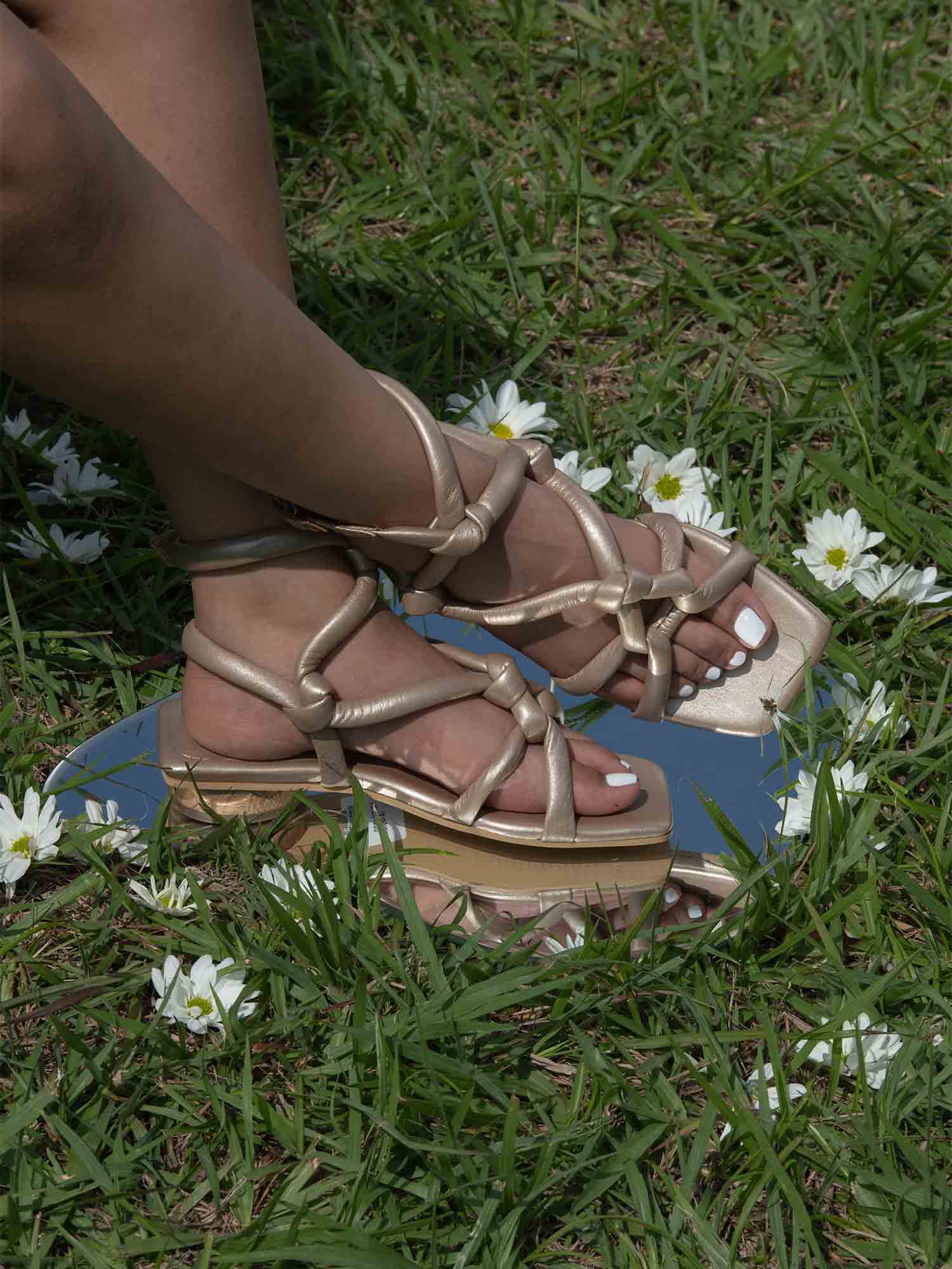 Detalle de los pies con la sandalia plana Duna dorada, capturando el brillo de los nudos artesanales y el mini tacón de madera bajo la luz natural