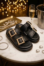 Sandalia modelo Gardenia en color negro, puestas en una mesa de mármol junto a varios accesorios de lujo destacando su elegancia