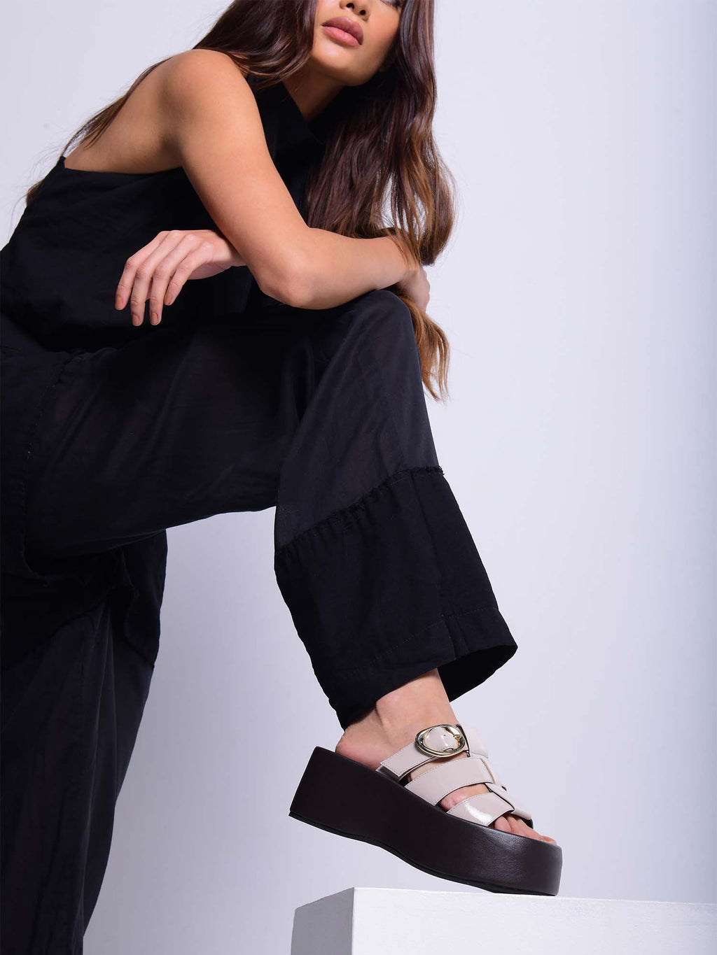 Modelo con enterizo negro posando con sandalias Alta beige sobre una plataforma, resaltando el diseño vanguardista y la altura del calzado