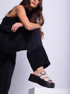 Modelo con enterizo negro posando con sandalias Alta beige sobre una plataforma, resaltando el diseño vanguardista y la altura del calzado