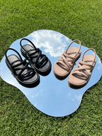 Imagen lifestyle del modelo de sandalias Tinna negras y frappé sobre un espejo en el césped, proyectando un estilo veraniego, moderno y versátil