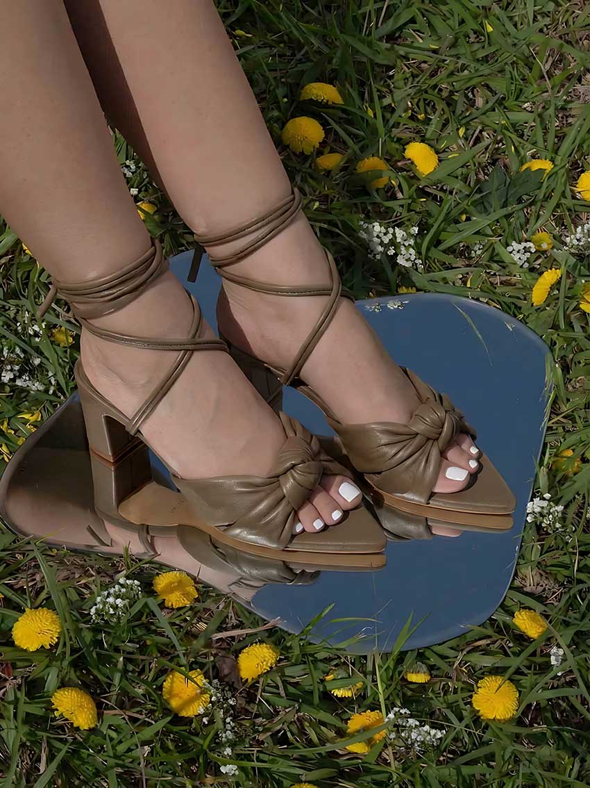 Imagen lifestyle de los pies luciendo los tacones Arden, capturando el efecto visual de las tiras amarradas al tobillo y el contraste del nudo acolchado.